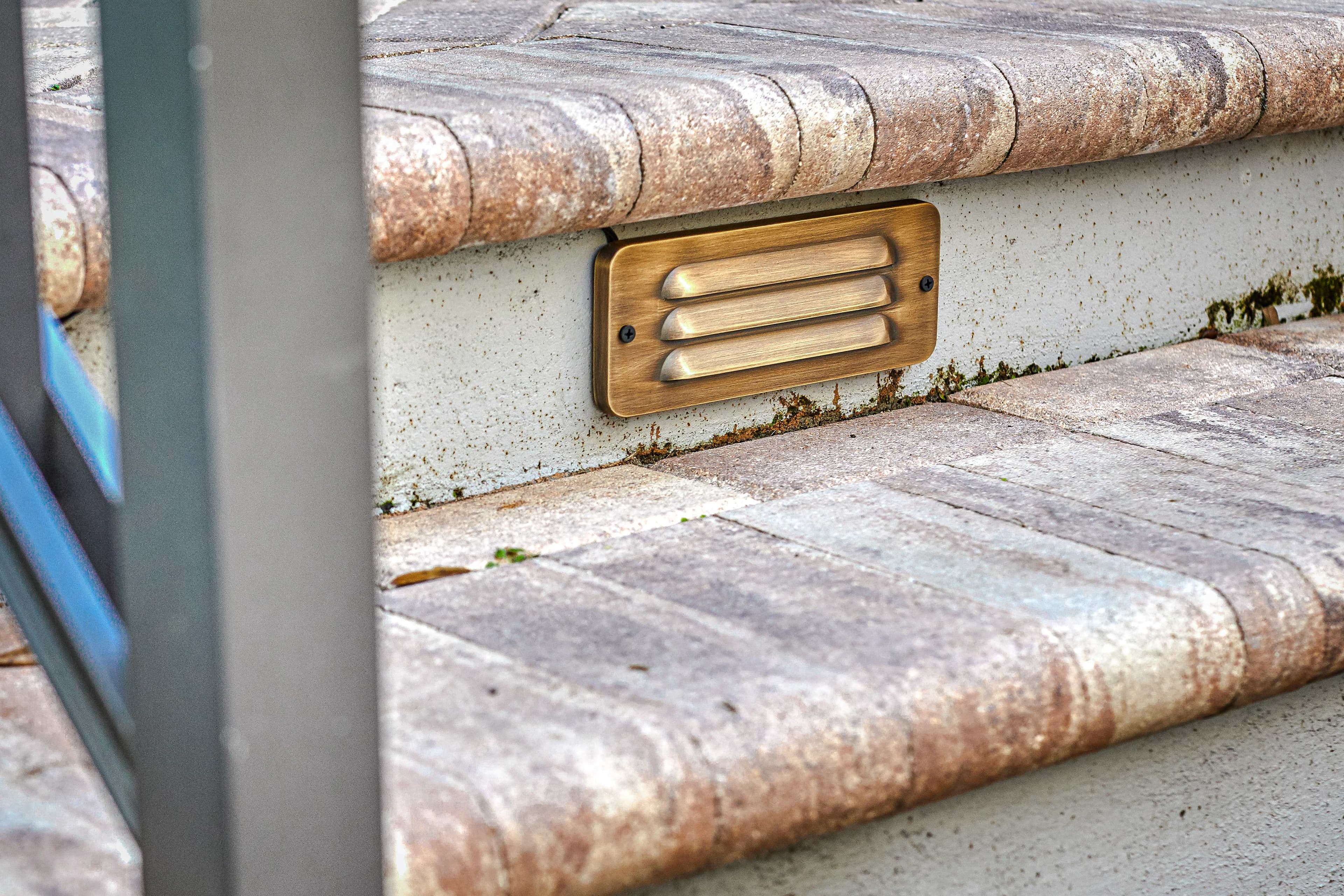 Close-up of brass step light fixture installed in stone wall near pavers showing quality craftsmanship