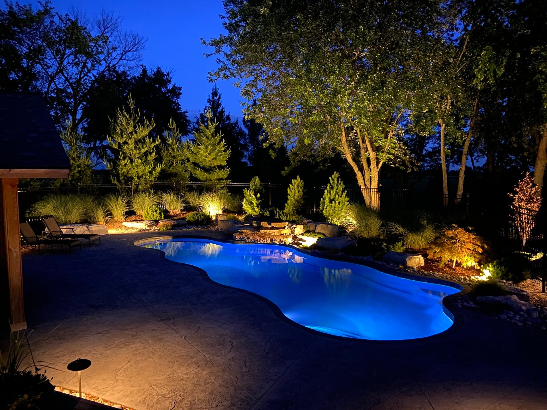 Beautiful pool at night with blue lighting surrounded by comprehensive landscape lighting including uplighting on trees, grasses, and rocks in Auburn Alabama