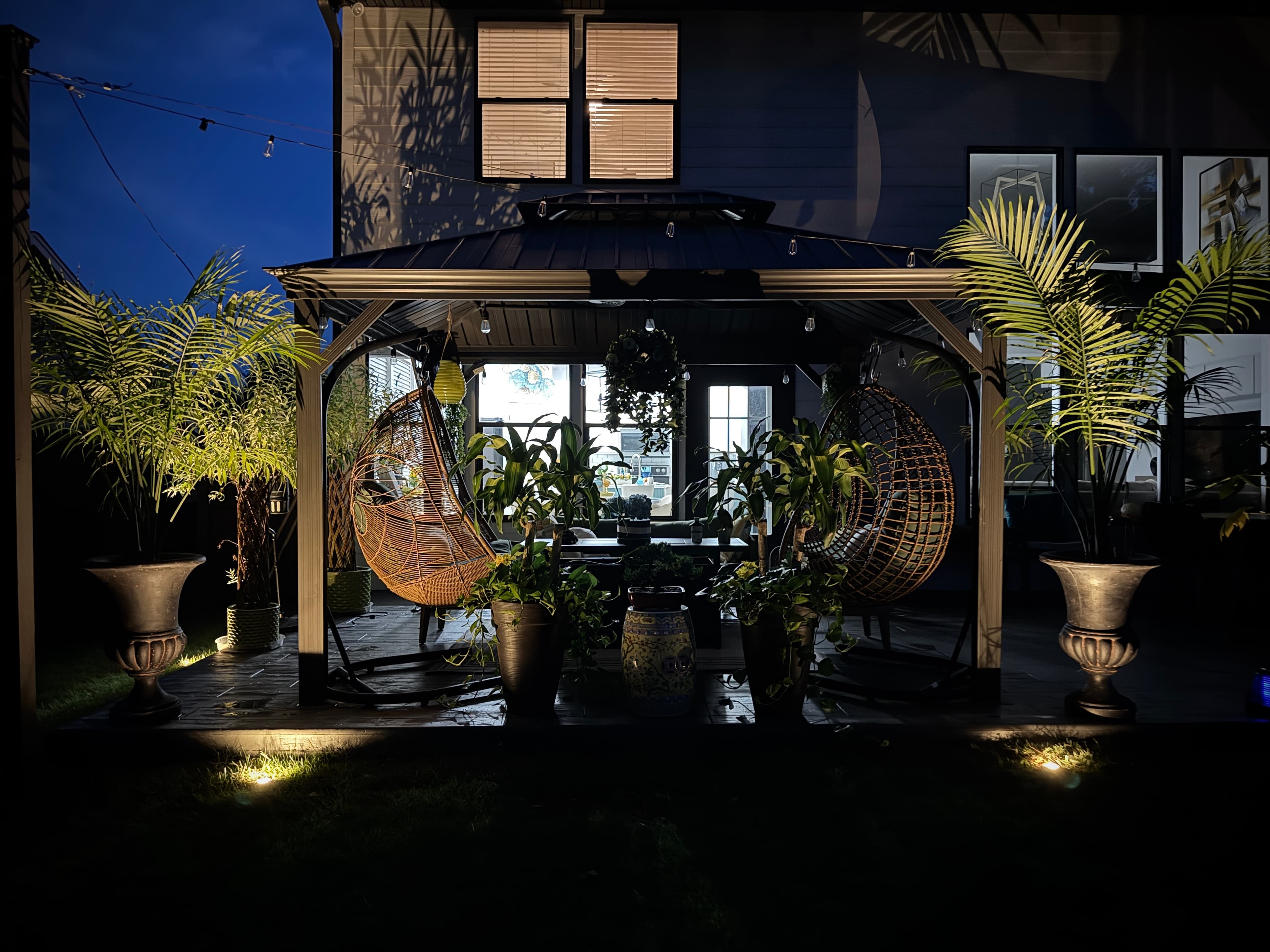 Outdoor patio at twilight with dual hanging wicker chairs, illuminated palm planters, and string lights overhead